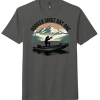 Charcoal perfect tri blend tee with 'Hooked Since Day One' fishing graphic and scenic mountain design.